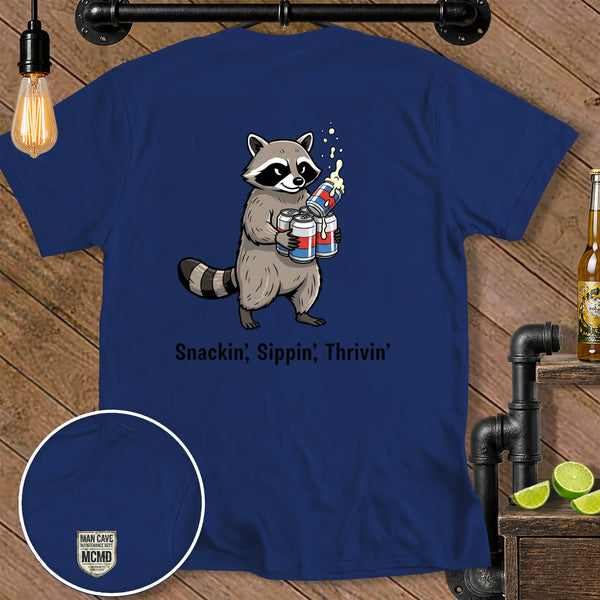 Navy t-shirt with raccoon holding beer cans and text "Snackin', Sippin', Thrivin'".