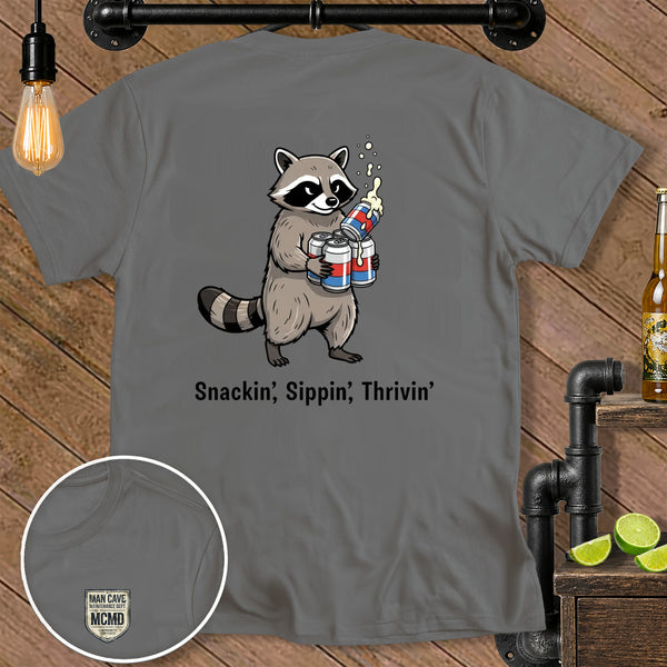 Gray t-shirt with cartoon raccoon holding cans and text "Snackin', Sippin', Thrivin'".
