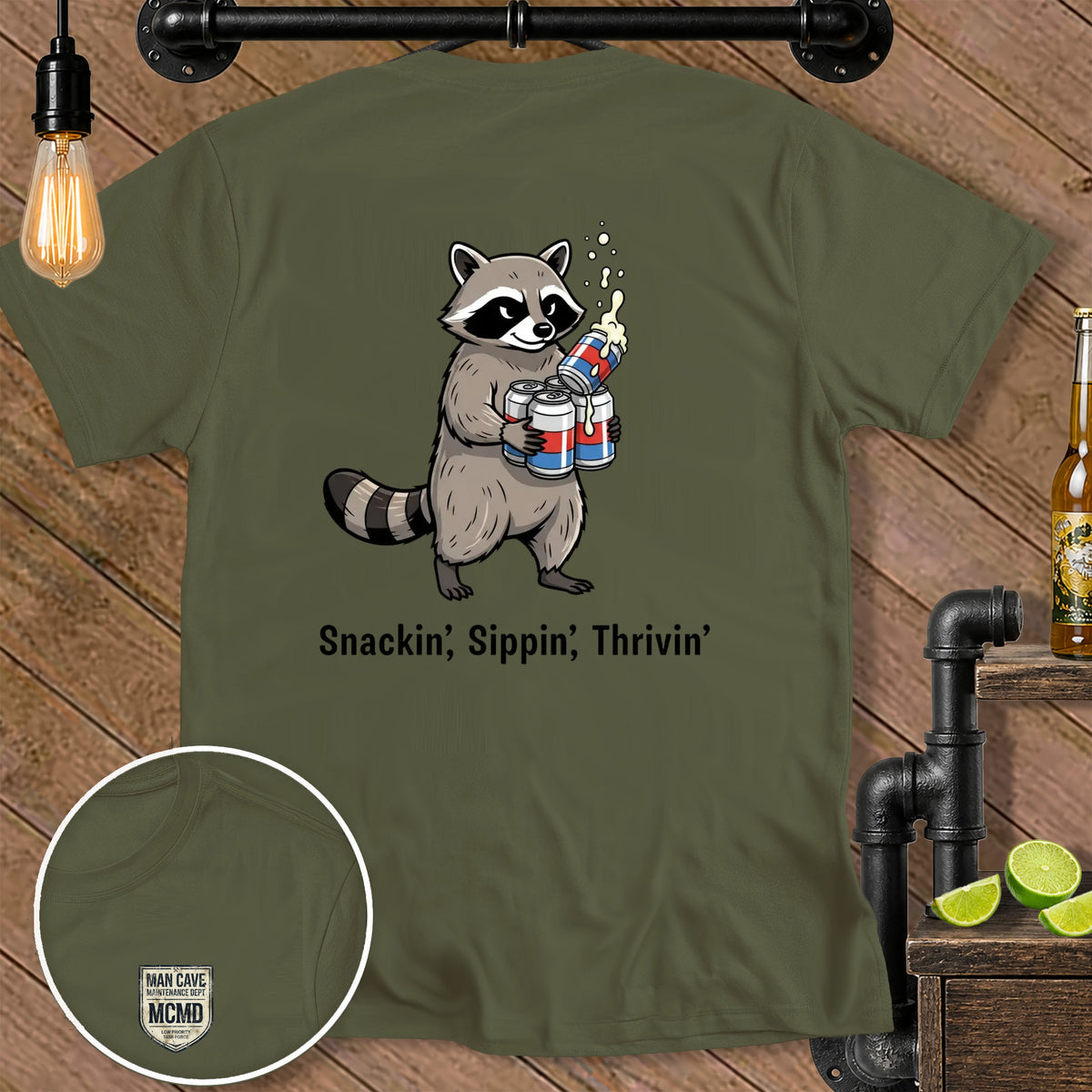 Olive green t-shirt with a raccoon holding cans and the text "Snackin', Sippin', Thrivin'".
